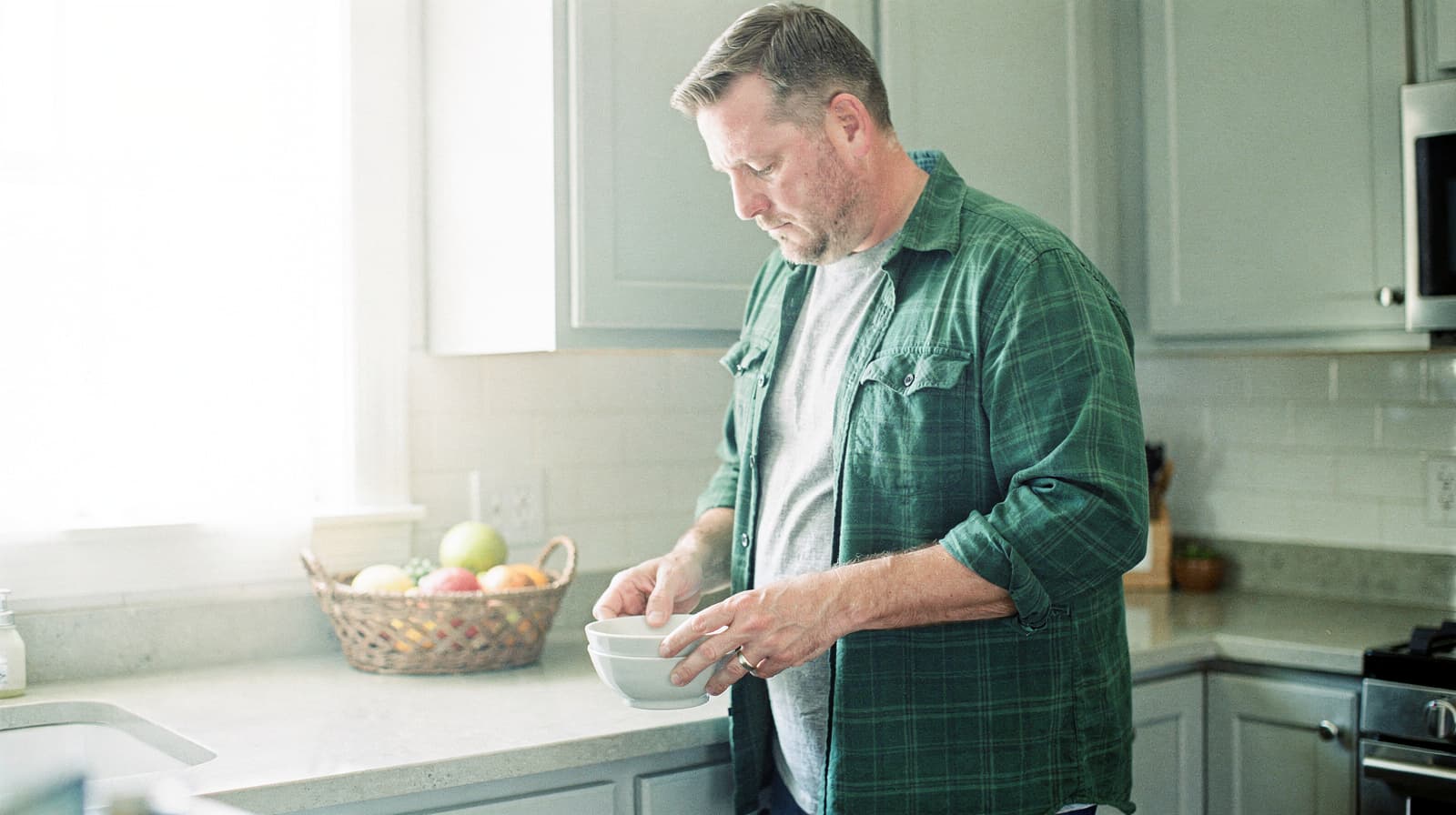 A man stands in a kitchen stirring food in a bowl near a basket of fruit on the counter.