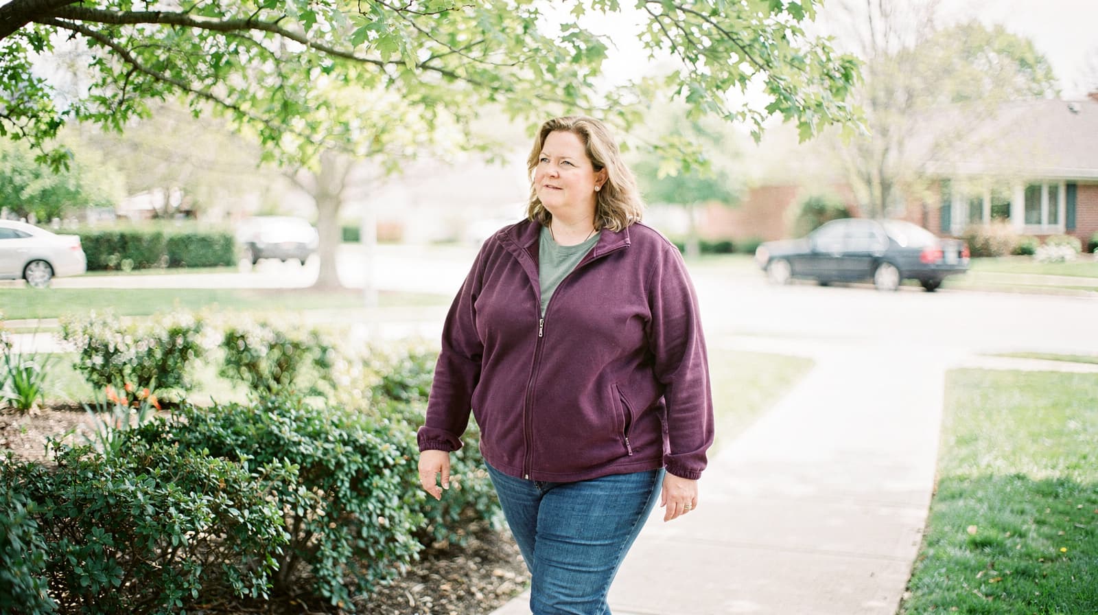 A woman in a purple jacket walks on a sidewalk in a suburban neighborhood with trees and parked cars.