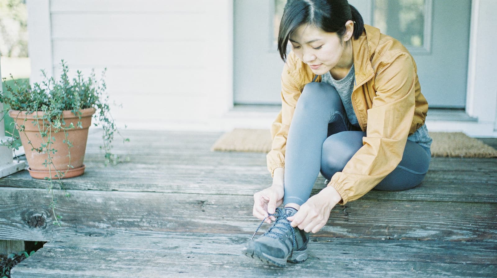A woman sitting on wooden steps ties her hiking boots next to a potted plant.
