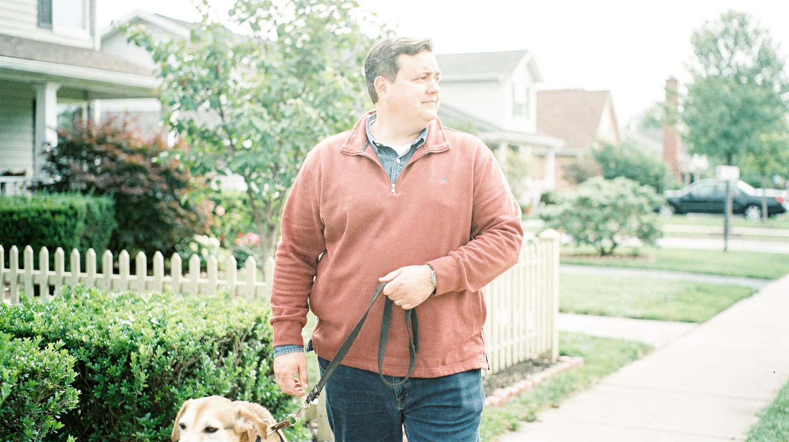 A man walks a dog on a sidewalk in a suburban neighborhood with houses and greenery.