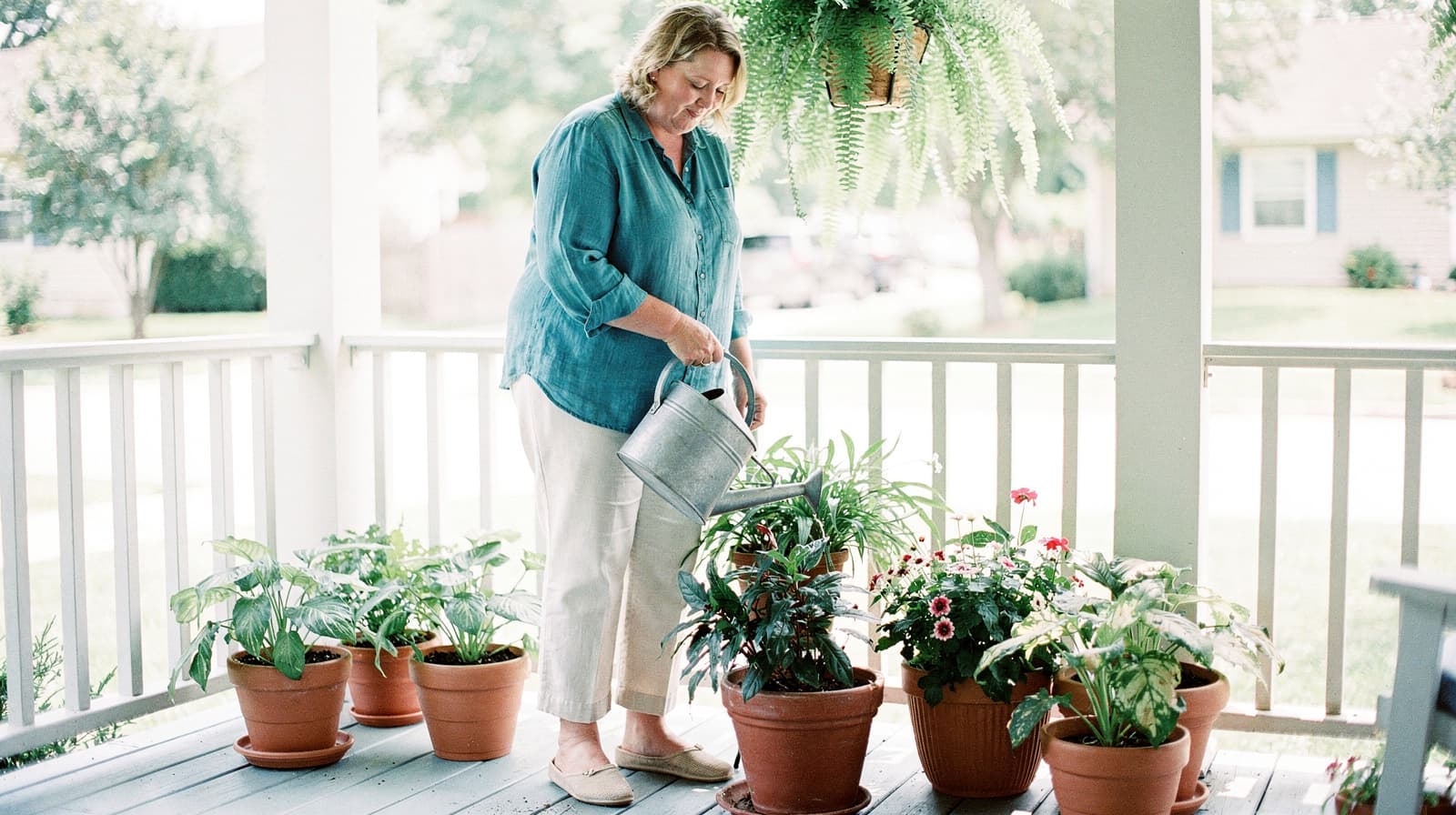 A woman waters potted plants on a porch with a hanging fern above.