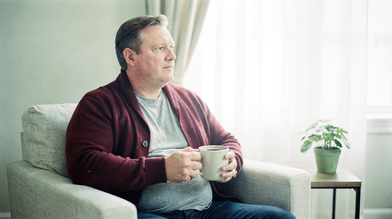 A man sits on a beige armchair holding a mug, with a small plant on a table by a window in the background.