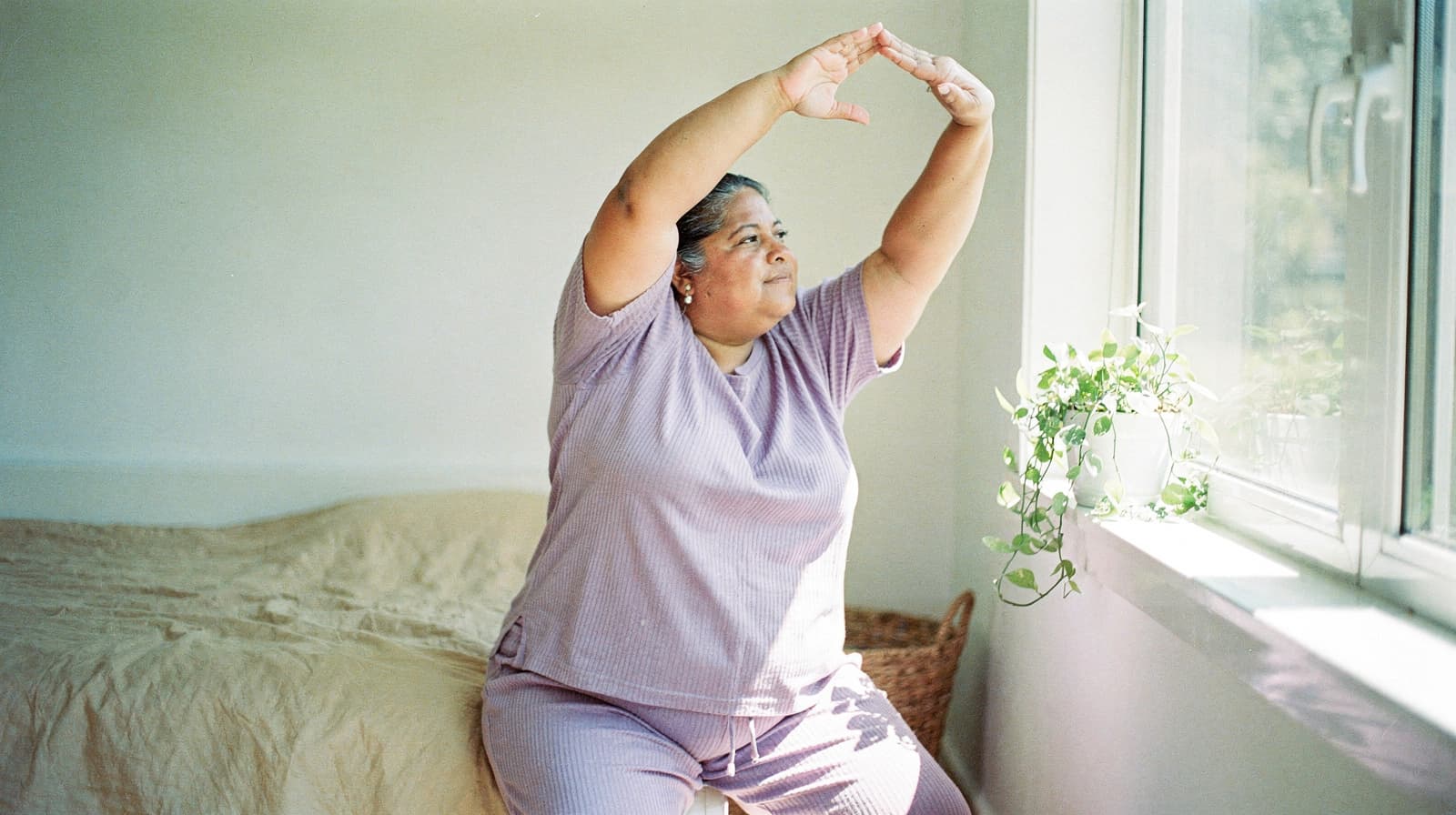 A woman in purple pajamas sits on a bed, stretching her arms toward a window with sunlight streaming in.