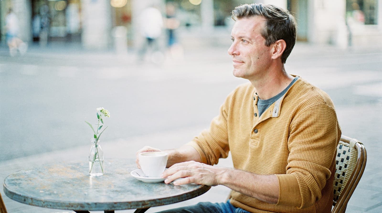 A man sits at an outdoor café table holding a cup, with a small vase of flowers in front of him.