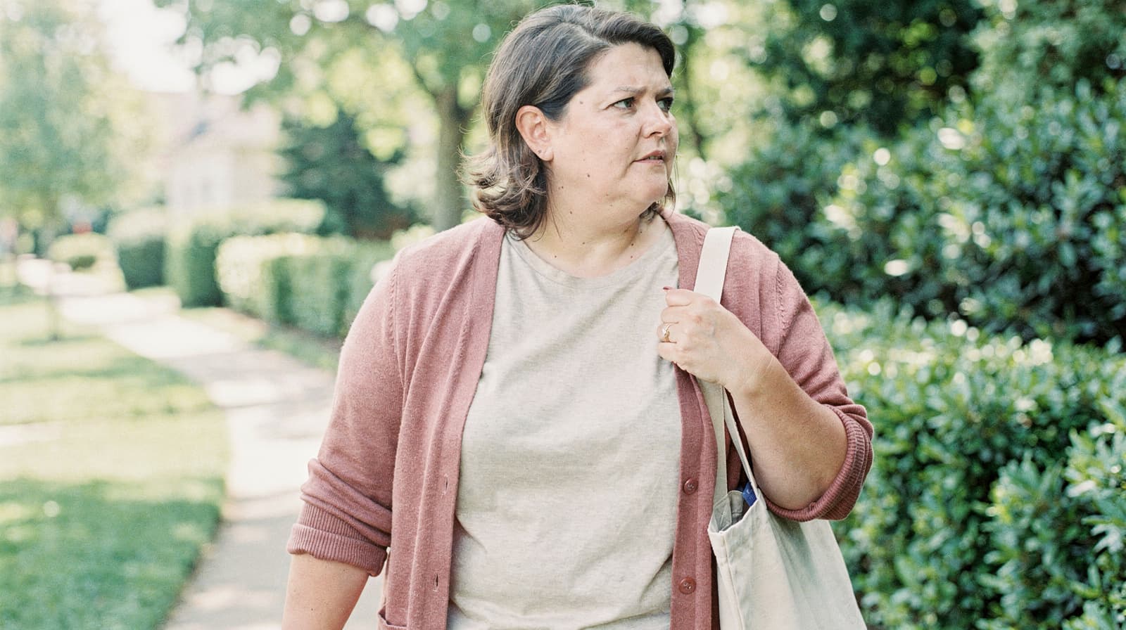 A woman walks on a sunny sidewalk carrying a tote bag, surrounded by greenery and trees.