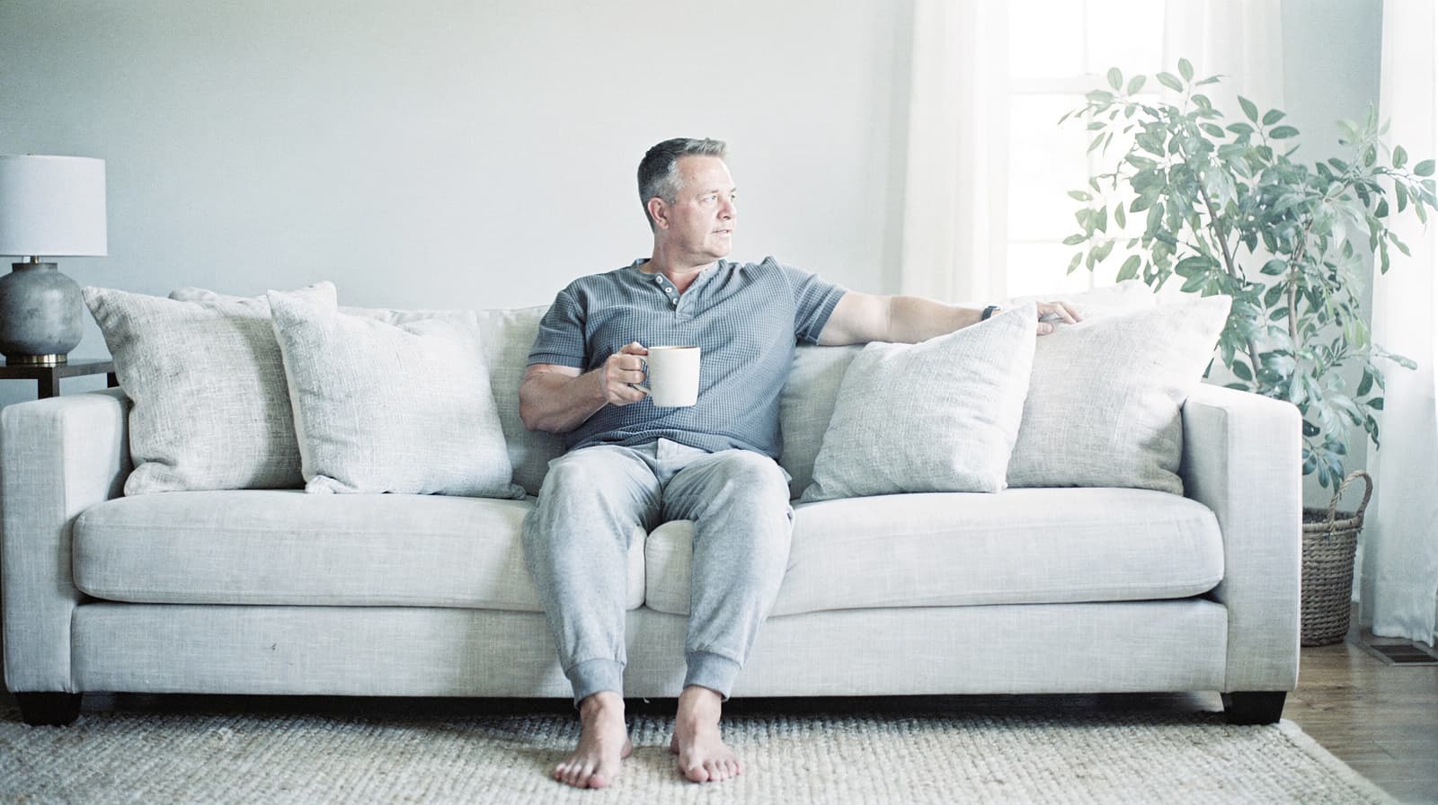 A man sits barefoot on a sofa holding a mug, looking out a window in a bright living room.
