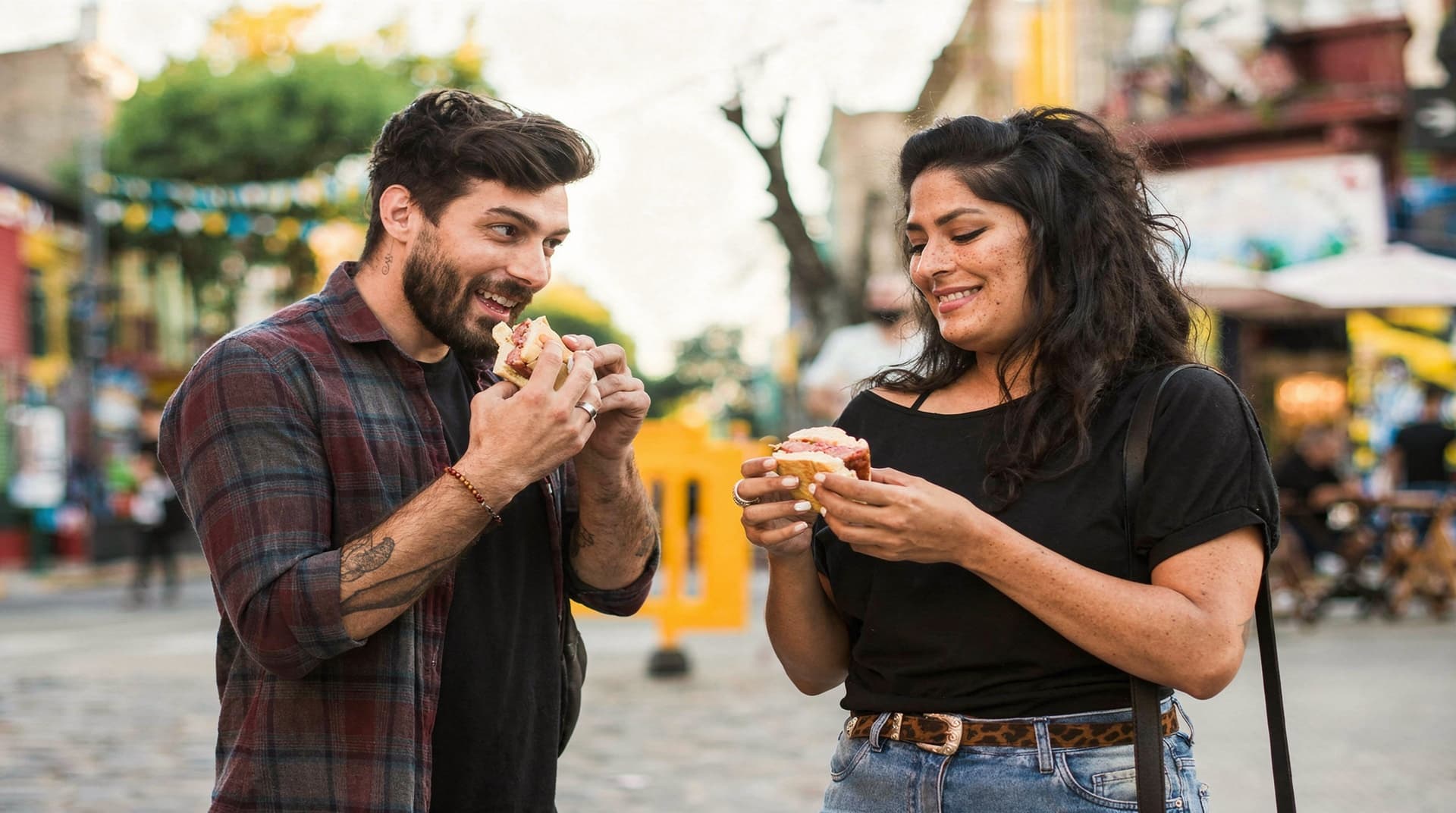 A man and a woman smile while eating sandwiches on a lively street with colorful buildings in the background.