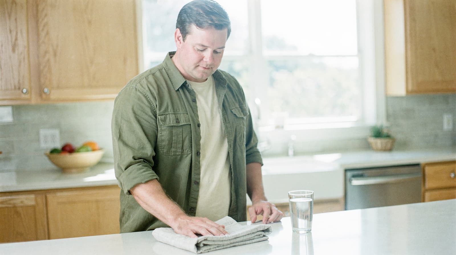 A man stands in a kitchen drying a glass with a towel near a countertop.