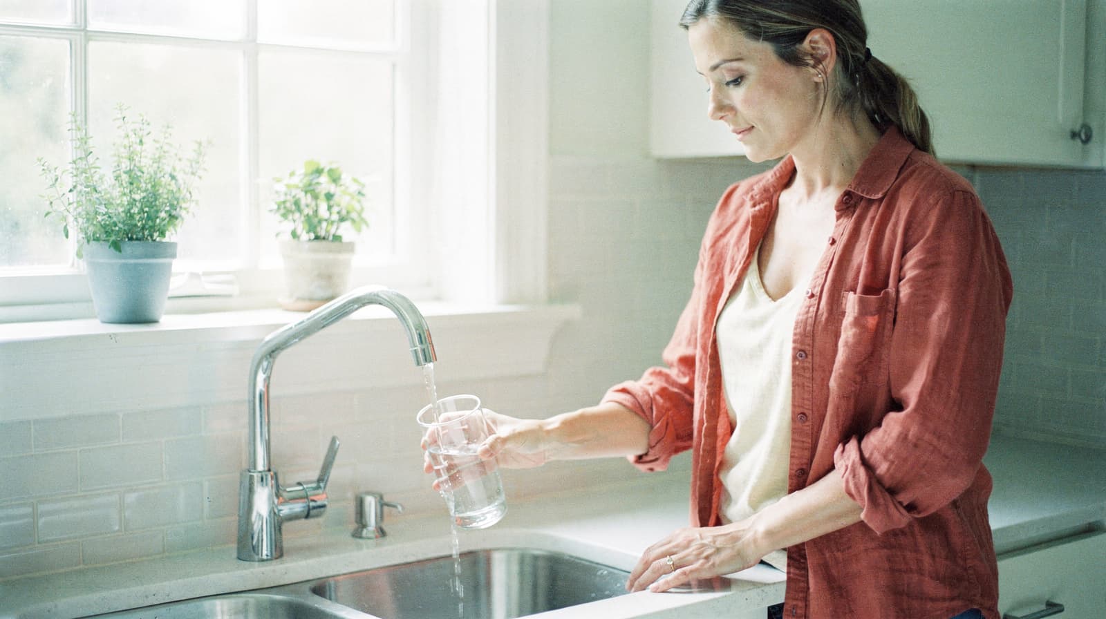 A woman fills a glass with water from a kitchen faucet near a window with potted plants.