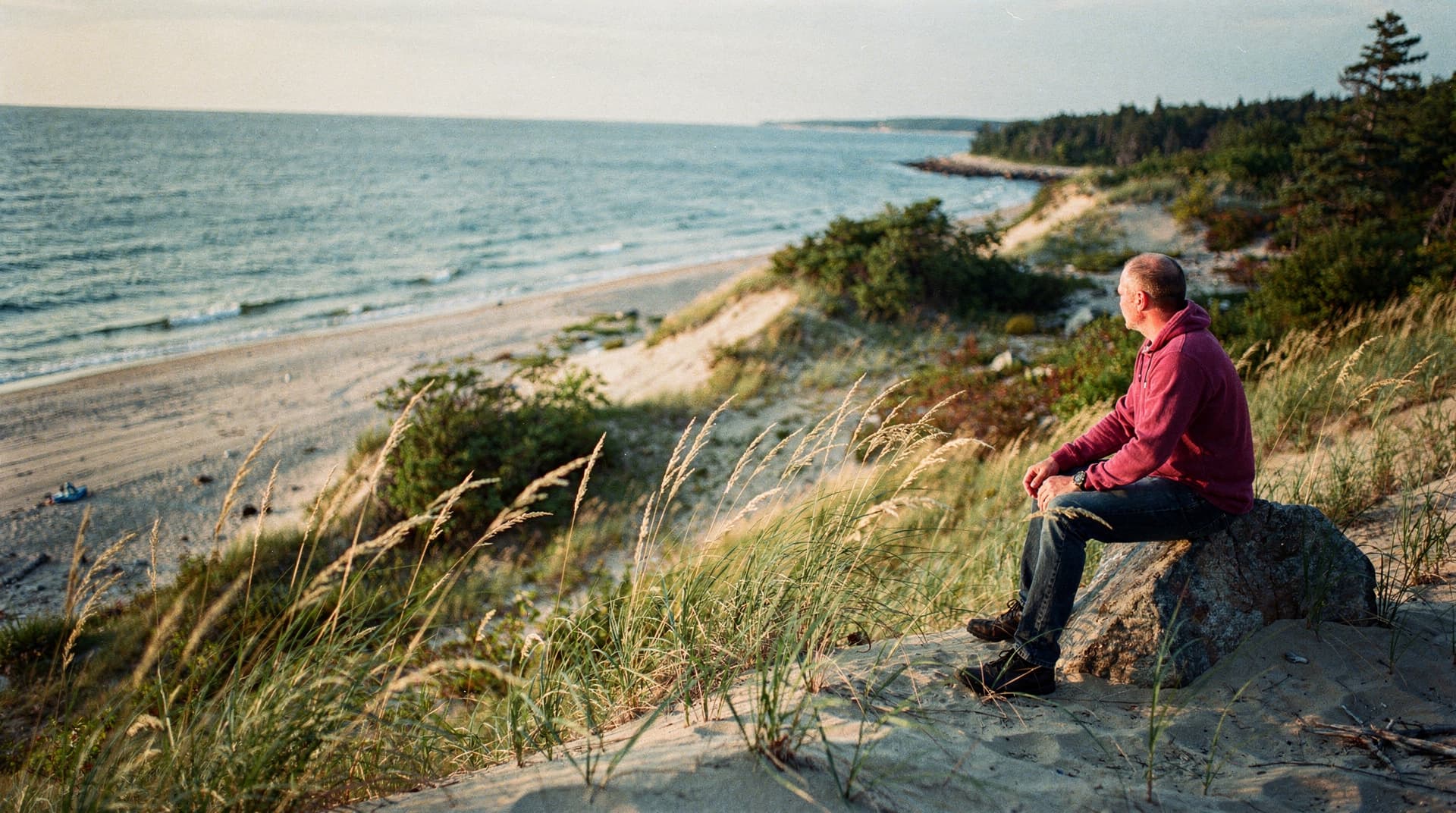 A man in a red hoodie sits on a rock overlooking a sandy beach and ocean with grassy dunes in the foreground.