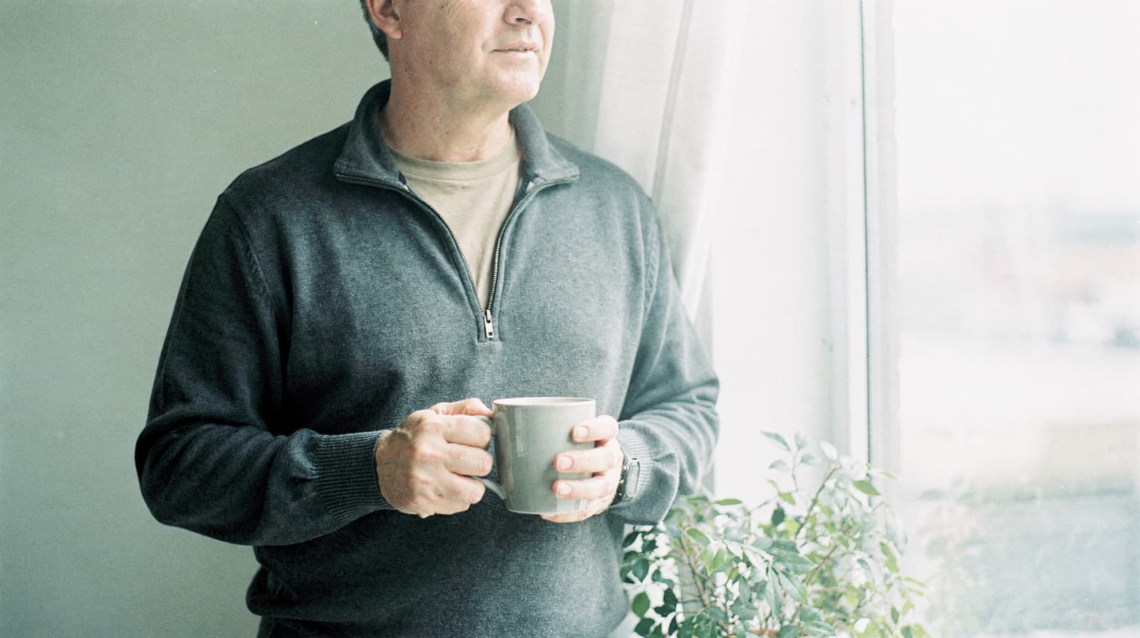 A man holding a mug stands by a window with a plant nearby.