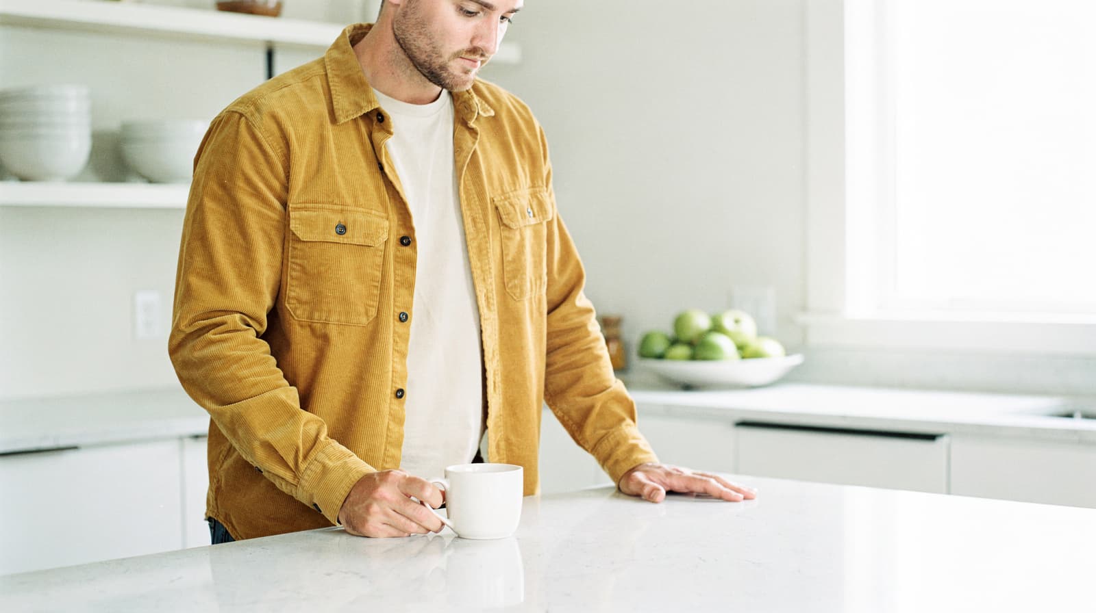 A man in a mustard jacket stands at a kitchen counter holding a white mug, with a bowl of green apples in the background.