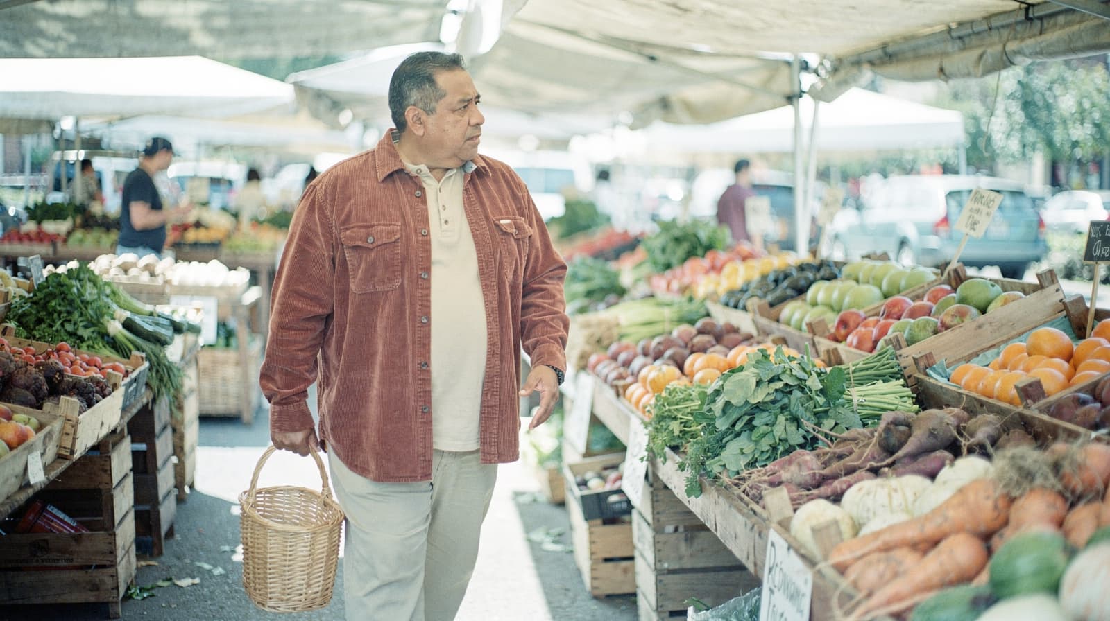 A man carrying a basket walks through a farmers market with various fruits and vegetables displayed on tables.