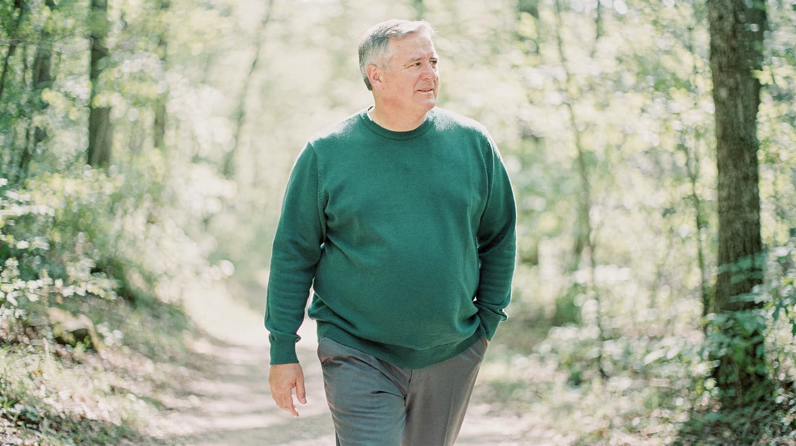 A man in a green sweater walks along a wooded path surrounded by trees and sunlight.