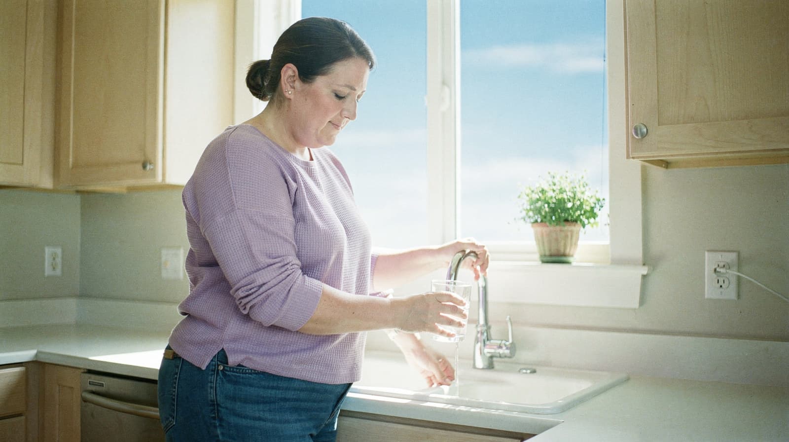 A woman fills a glass with water at a kitchen sink, with sunlight streaming through a window.