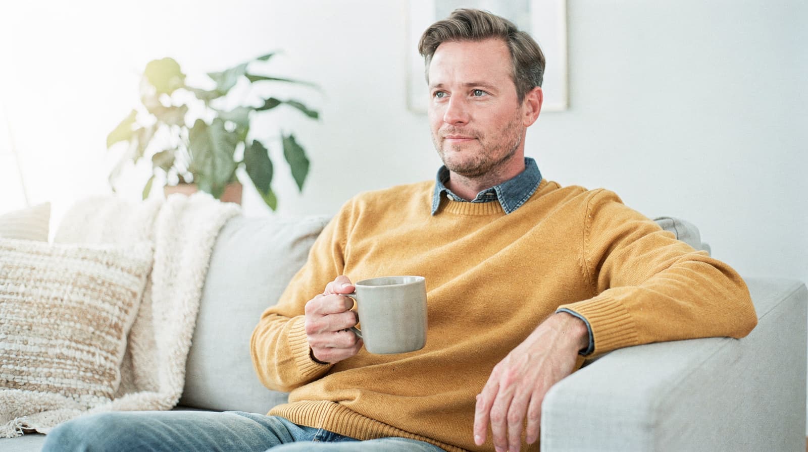 A man in a yellow sweater sits on a couch holding a mug, with a plant and a blanket in the background.