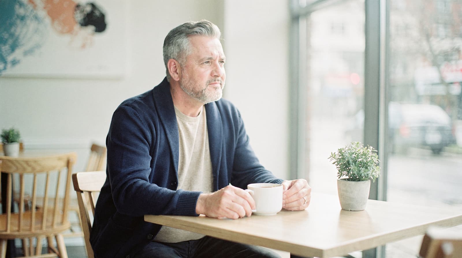 A man sits at a wooden table in a cafe, holding a cup and looking out the window.