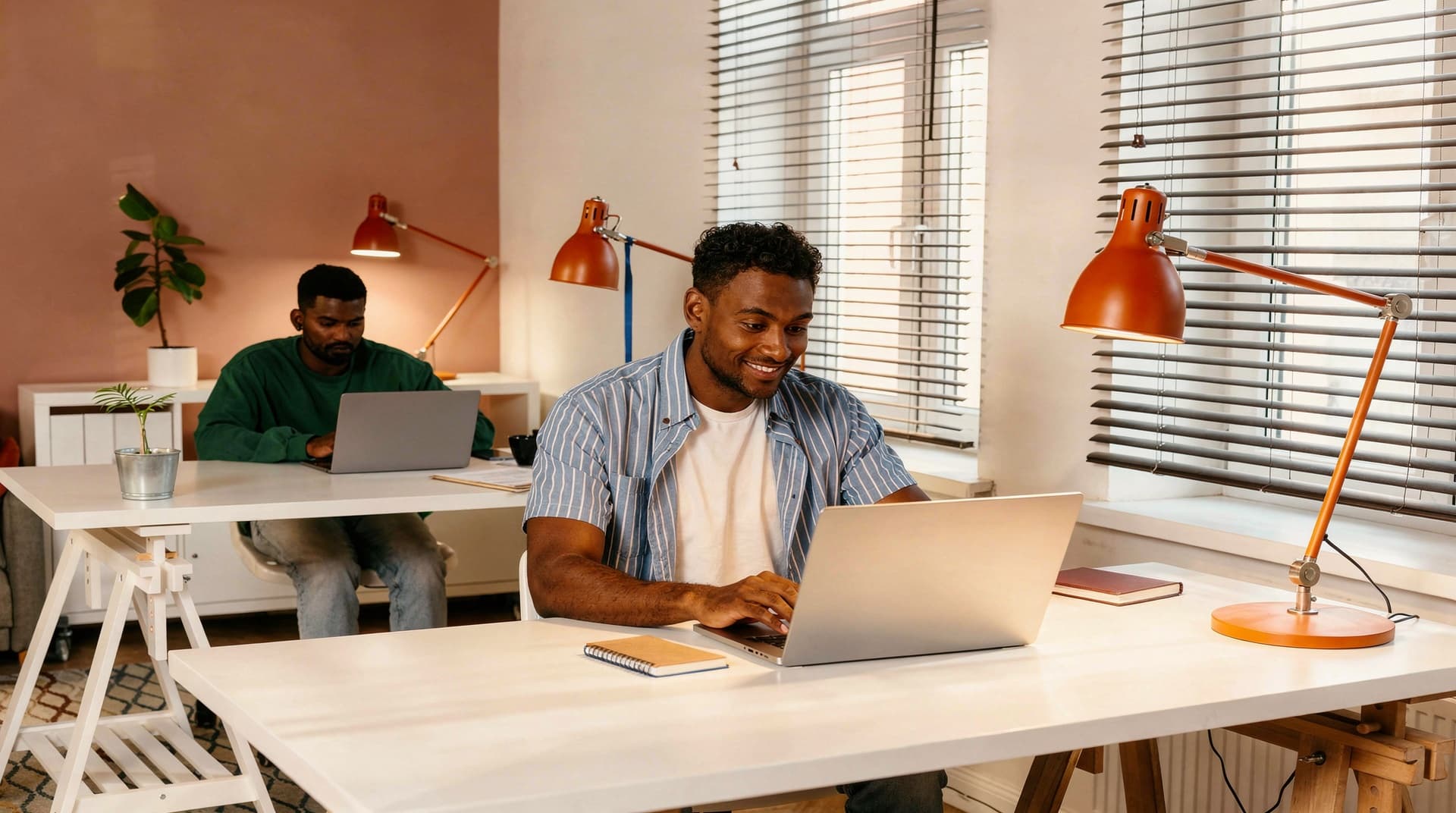 Two men work on laptops at separate desks in a bright office with large windows and orange desk lamps.