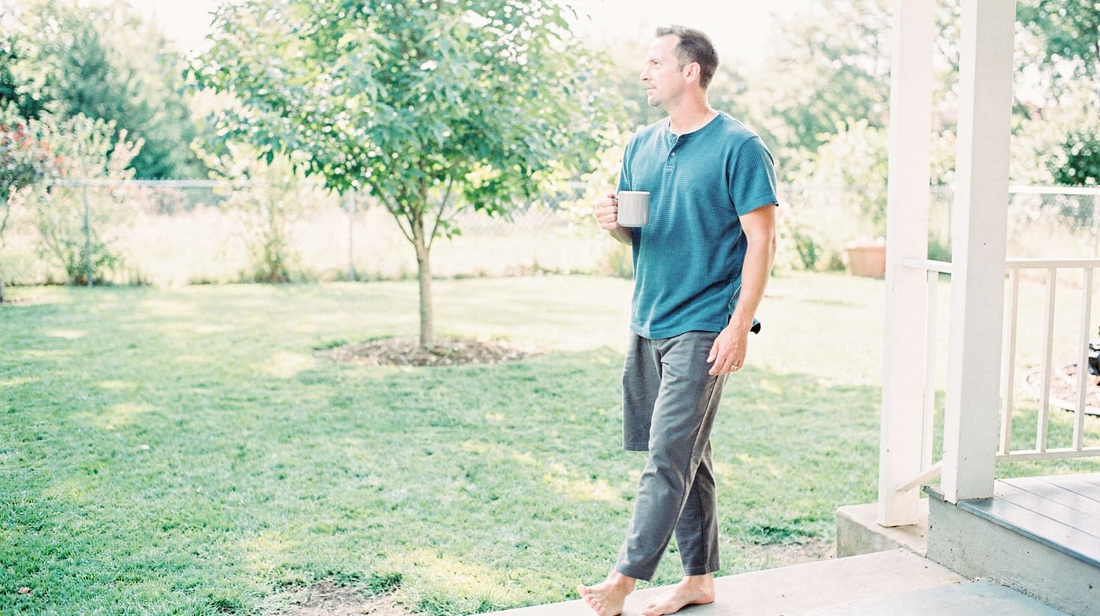 A man stands barefoot on a porch holding a mug, looking out at a grassy backyard with a tree.