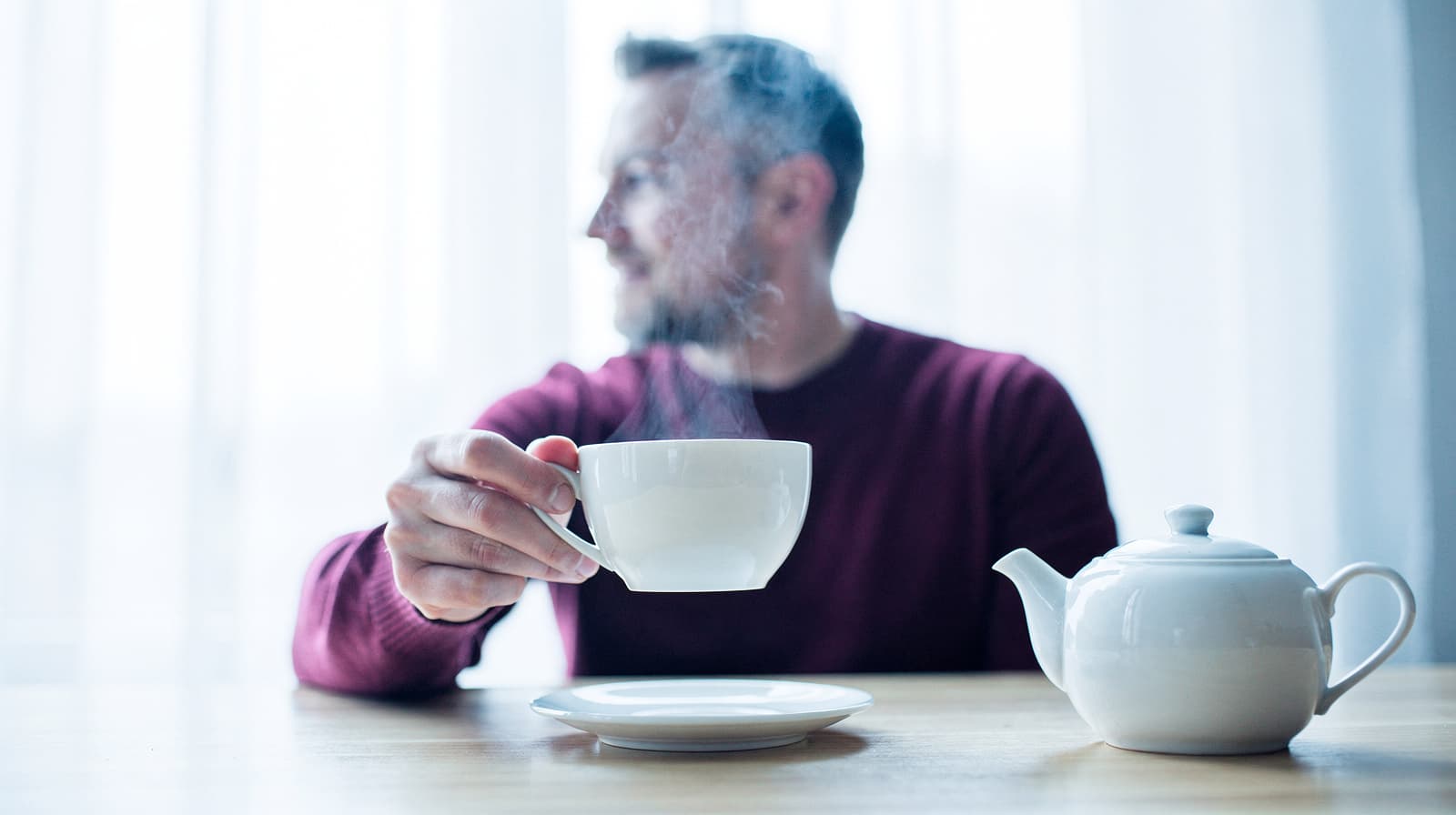 A man in a red sweater holds a steaming cup at a table with a white teapot.