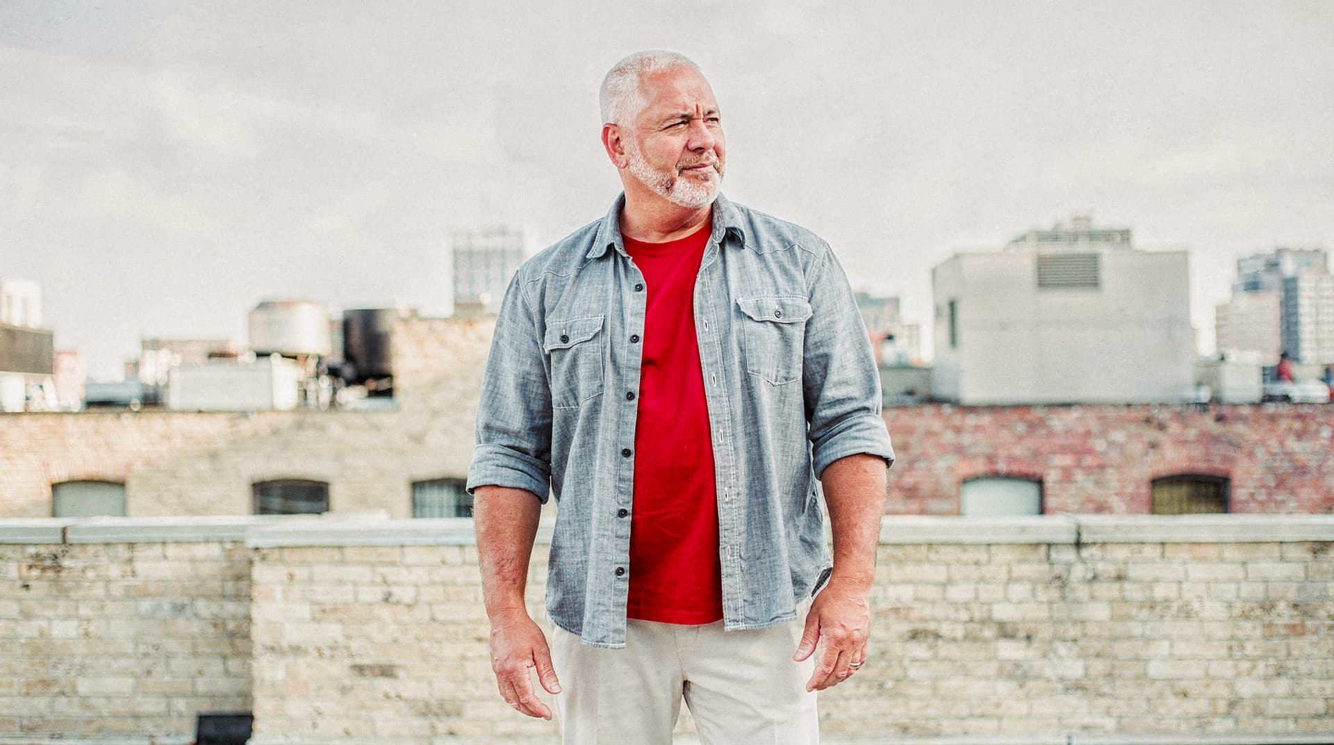 A man with a beard stands on a rooftop with a cityscape in the background.