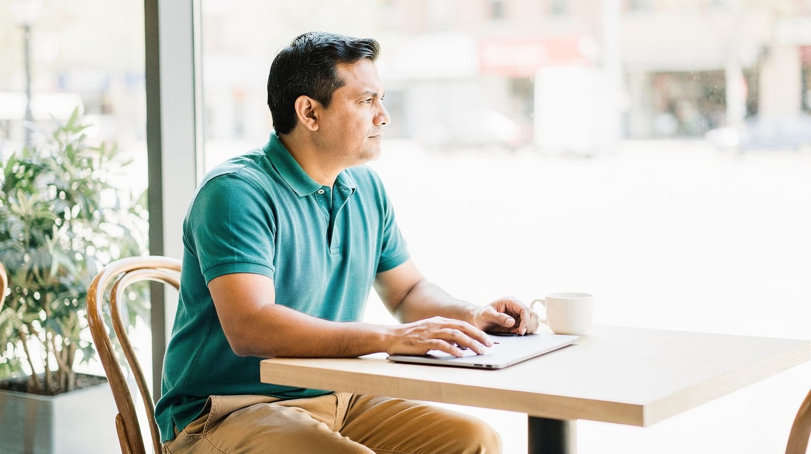 A man in a teal shirt sits at a table with a laptop and a cup, looking out a window in a bright cafe.