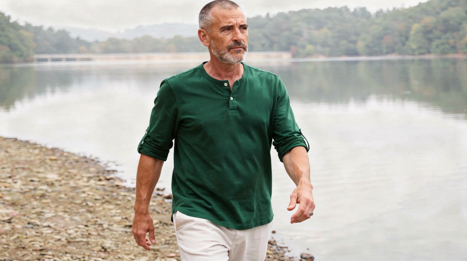A man in a green shirt walks along a rocky lakeshore with trees and hills in the background.