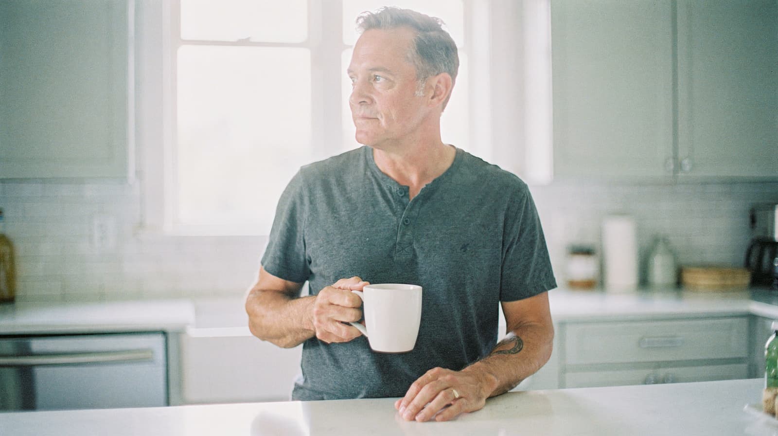 A man holding a mug stands in a kitchen, looking out the window.