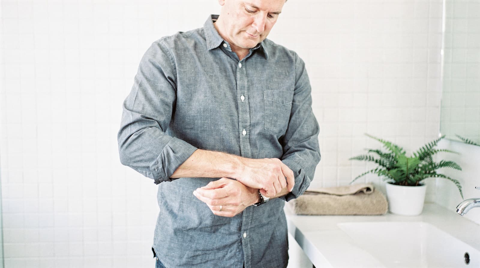 A man in a gray shirt stands in a bathroom adjusting his watch near a sink with a potted plant.