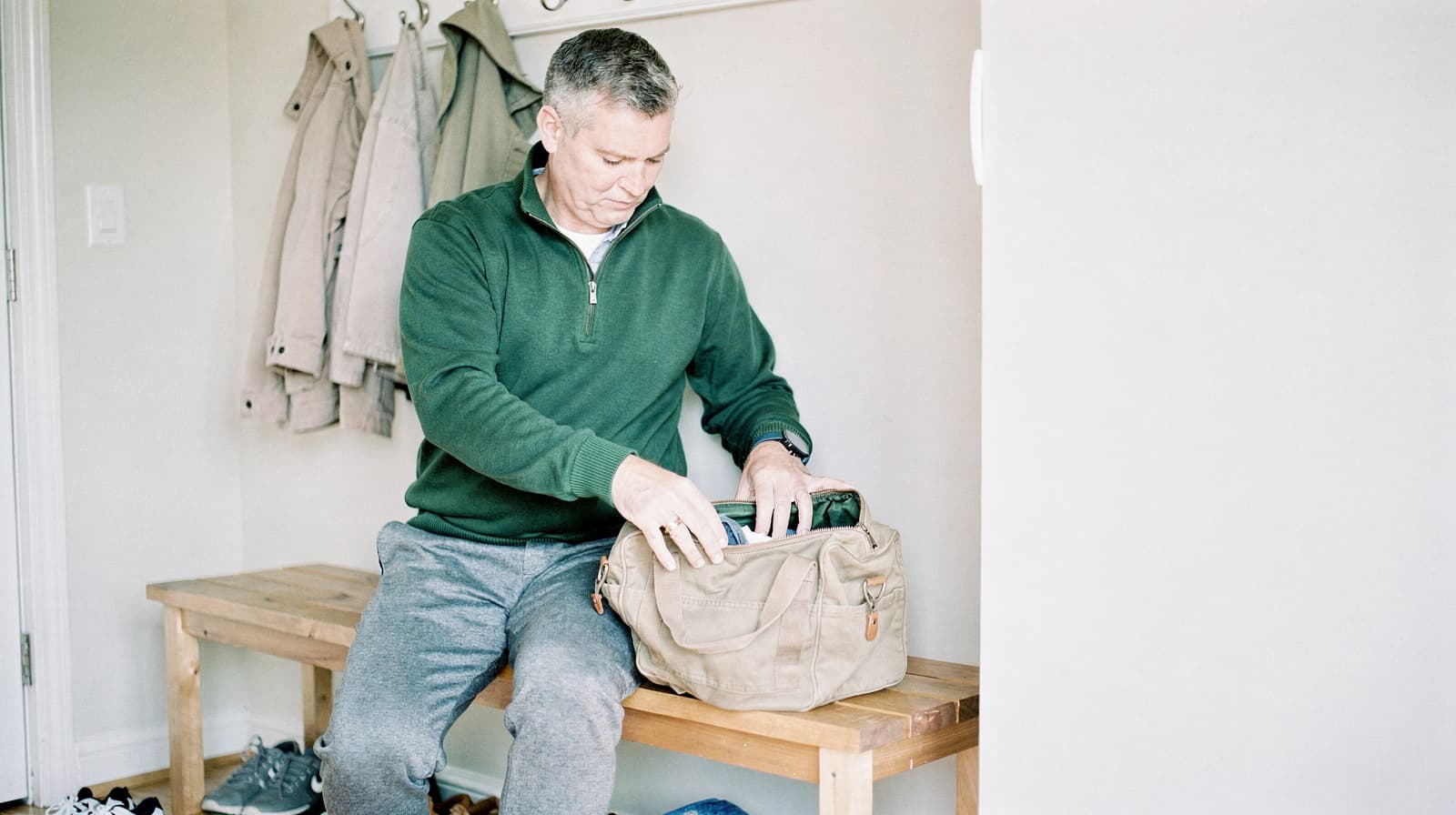 A man in a green sweater sits on a wooden bench, packing items into a beige bag in a hallway.