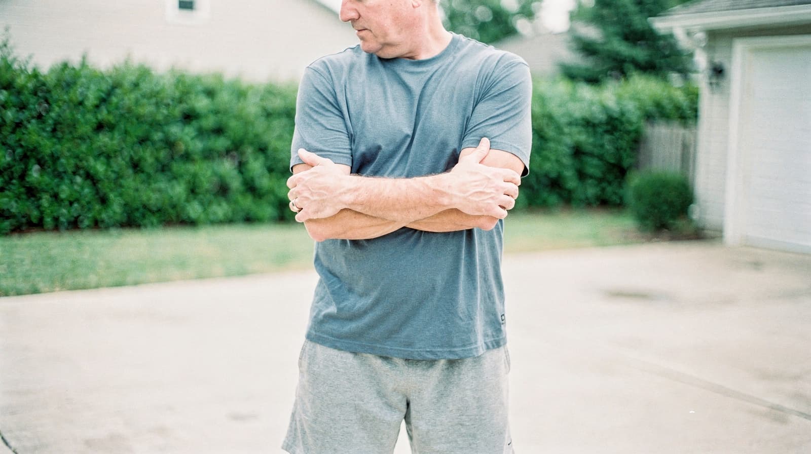 A man in a gray t-shirt and shorts stands with arms crossed in a driveway.