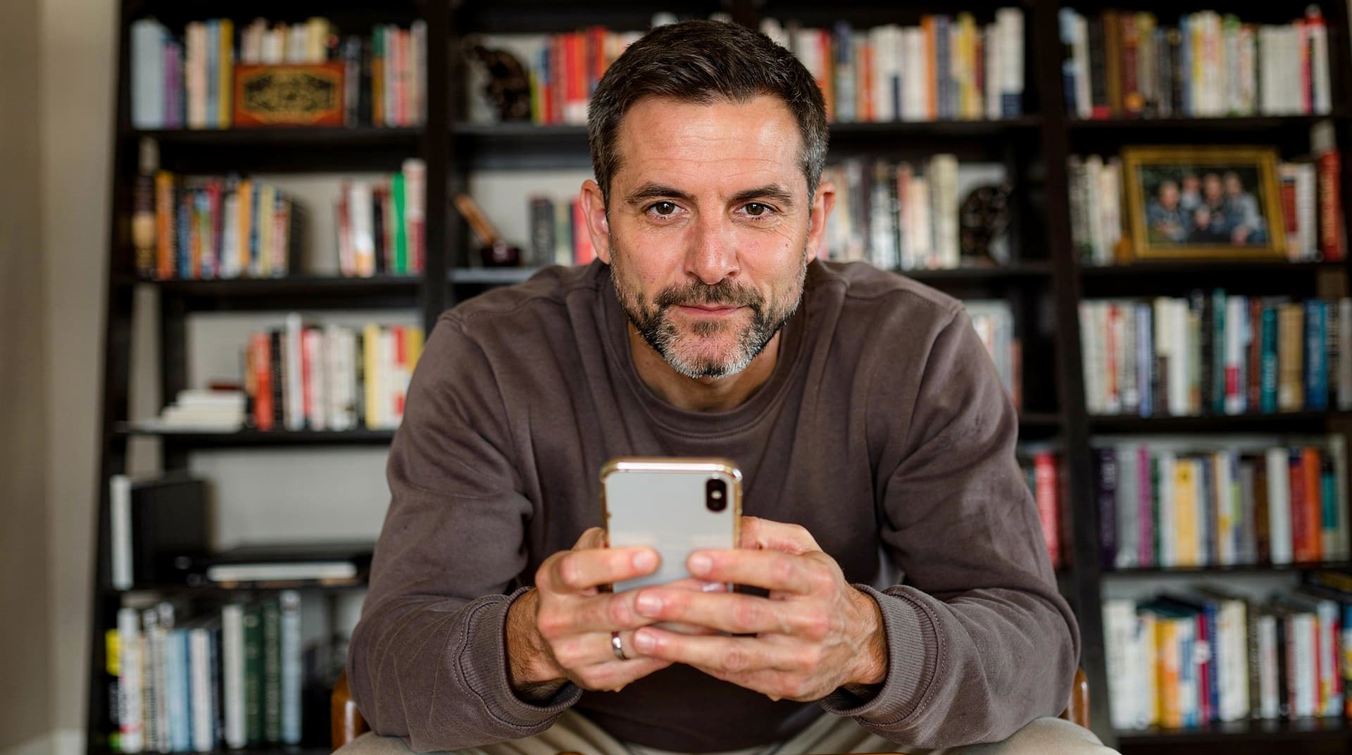 A man in a brown sweater sits in front of a bookshelf, holding a smartphone and looking at the camera.