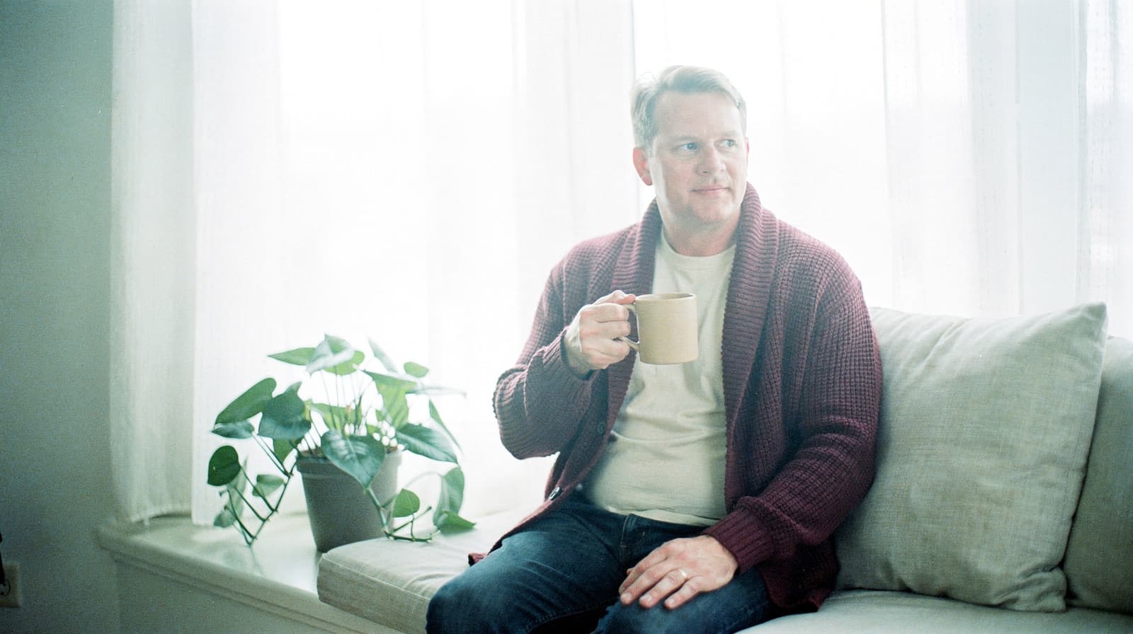 A man sits on a window seat holding a mug, with a potted plant beside him.