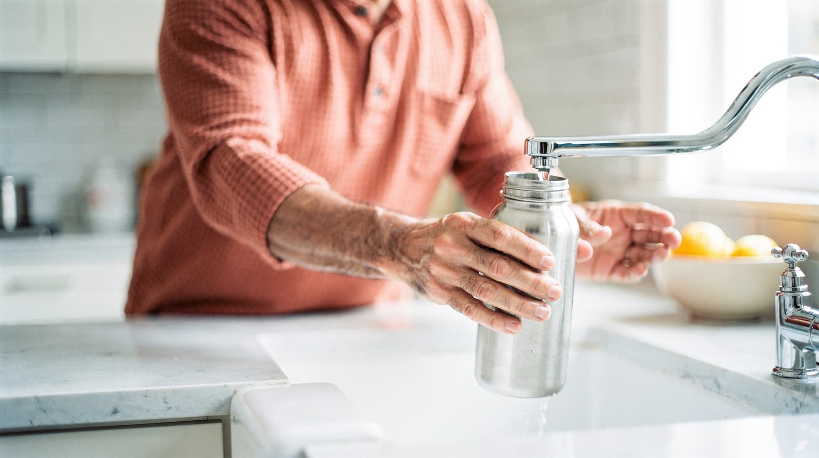A person fills a metal water bottle from a kitchen faucet near a bowl of lemons.