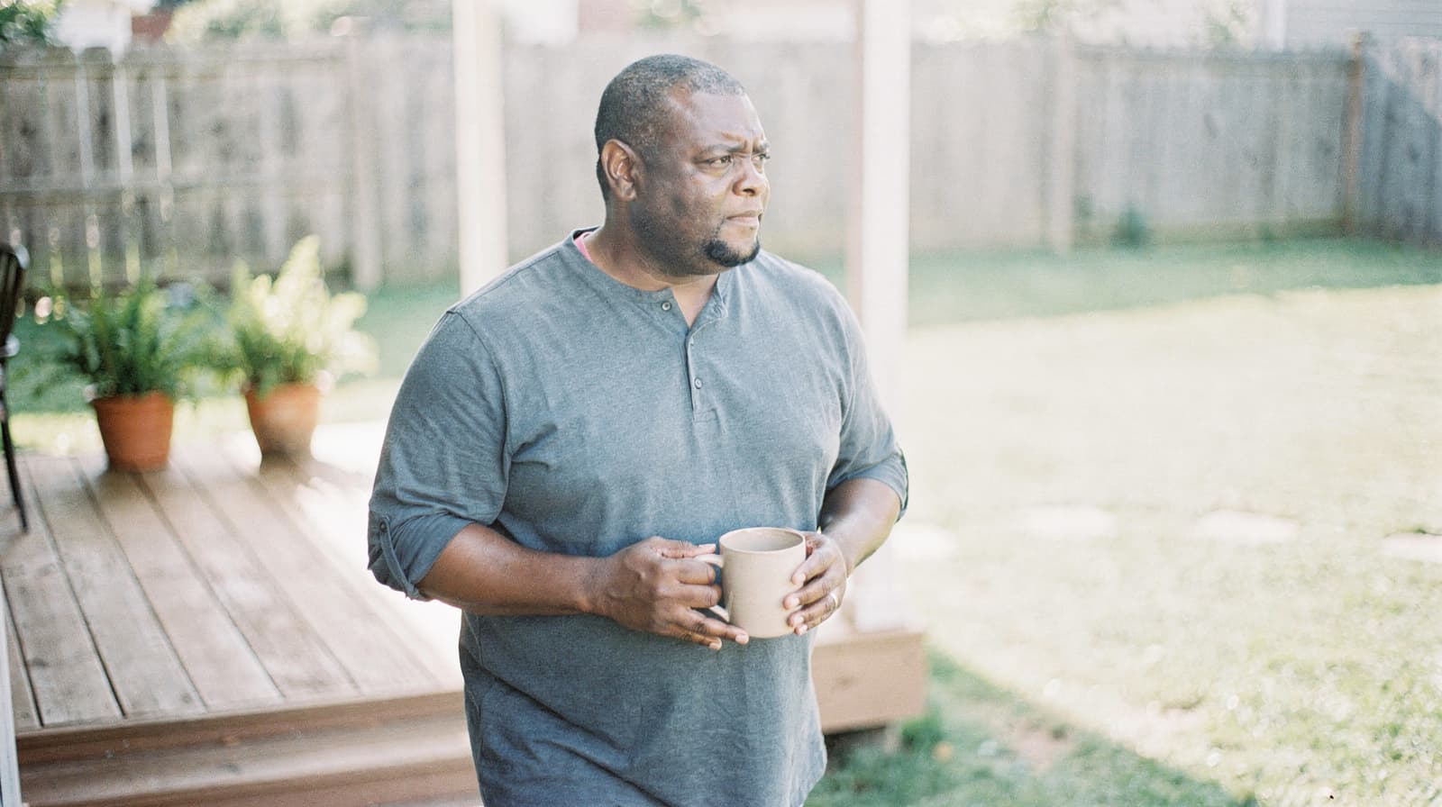 A man holding a mug stands on a porch looking out at a grassy backyard.