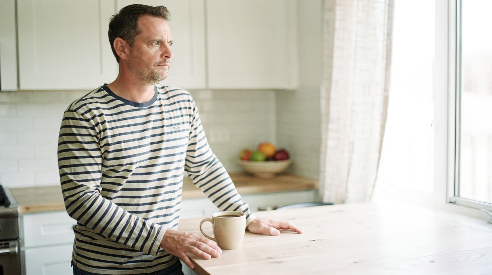A man in a striped shirt stands at a wooden table holding a mug, looking out a window in a bright kitchen.