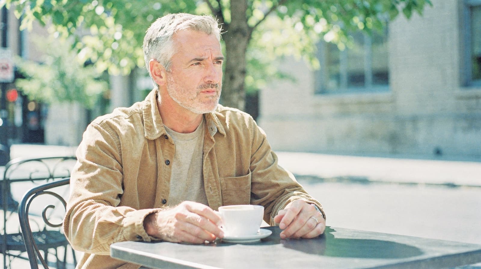 A man sits at an outdoor café table holding a cup, with trees and a building in the background.