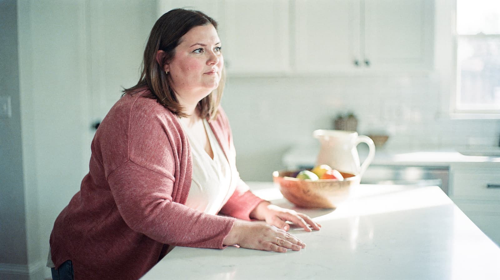 A woman in a pink cardigan leans on a kitchen counter with a bowl of fruit and a pitcher in the background.