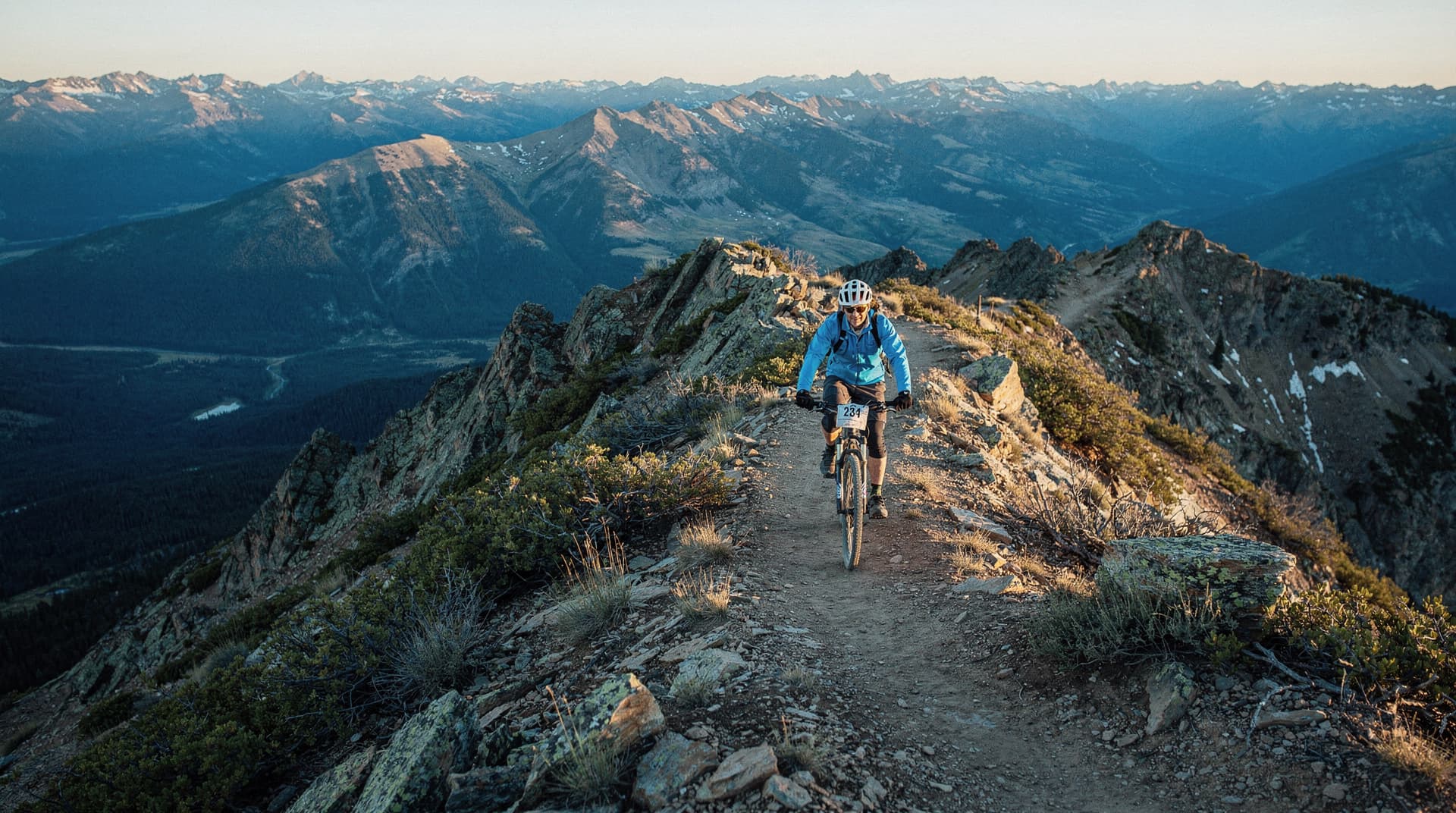 A person wearing a helmet rides a mountain bike along a narrow trail on a rocky ridge with distant mountain views.