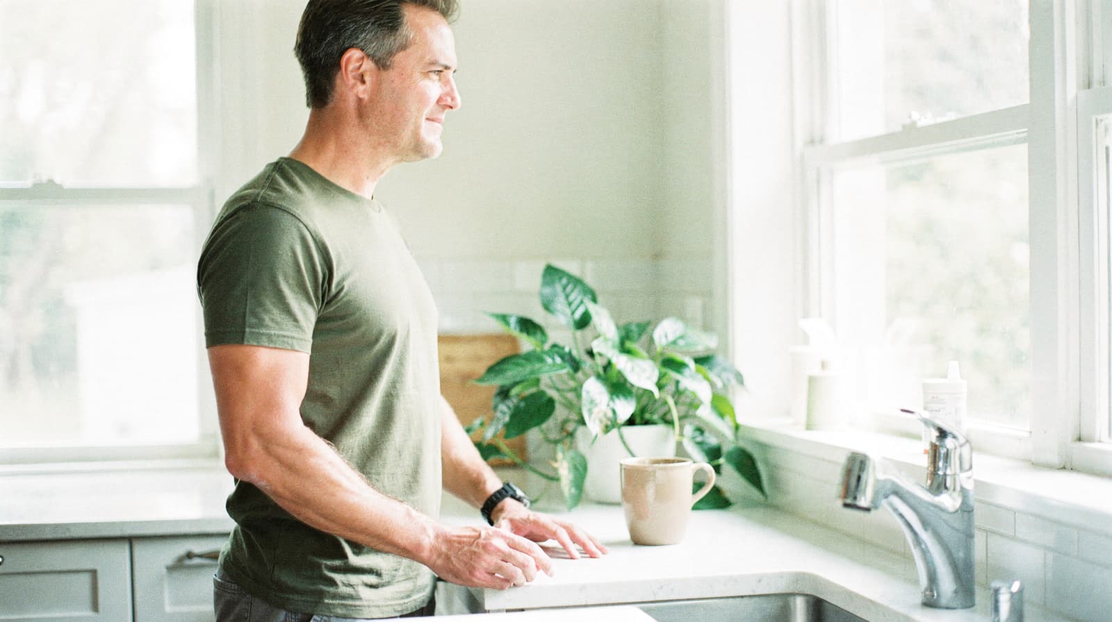 A man stands in a kitchen looking out a window with a plant and mug on the counter.