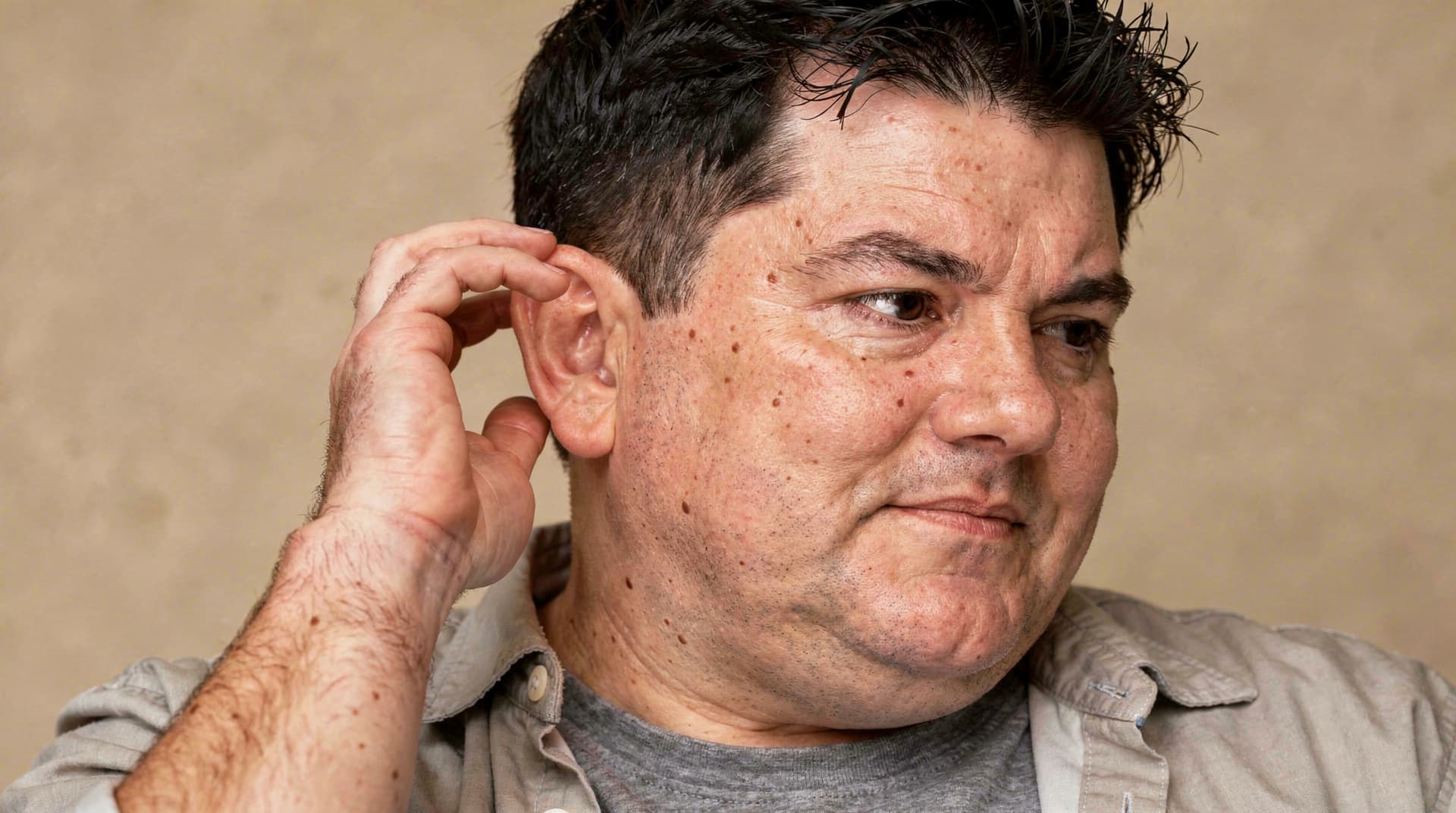 A man with short dark hair touches his ear while looking to the side against a plain background.