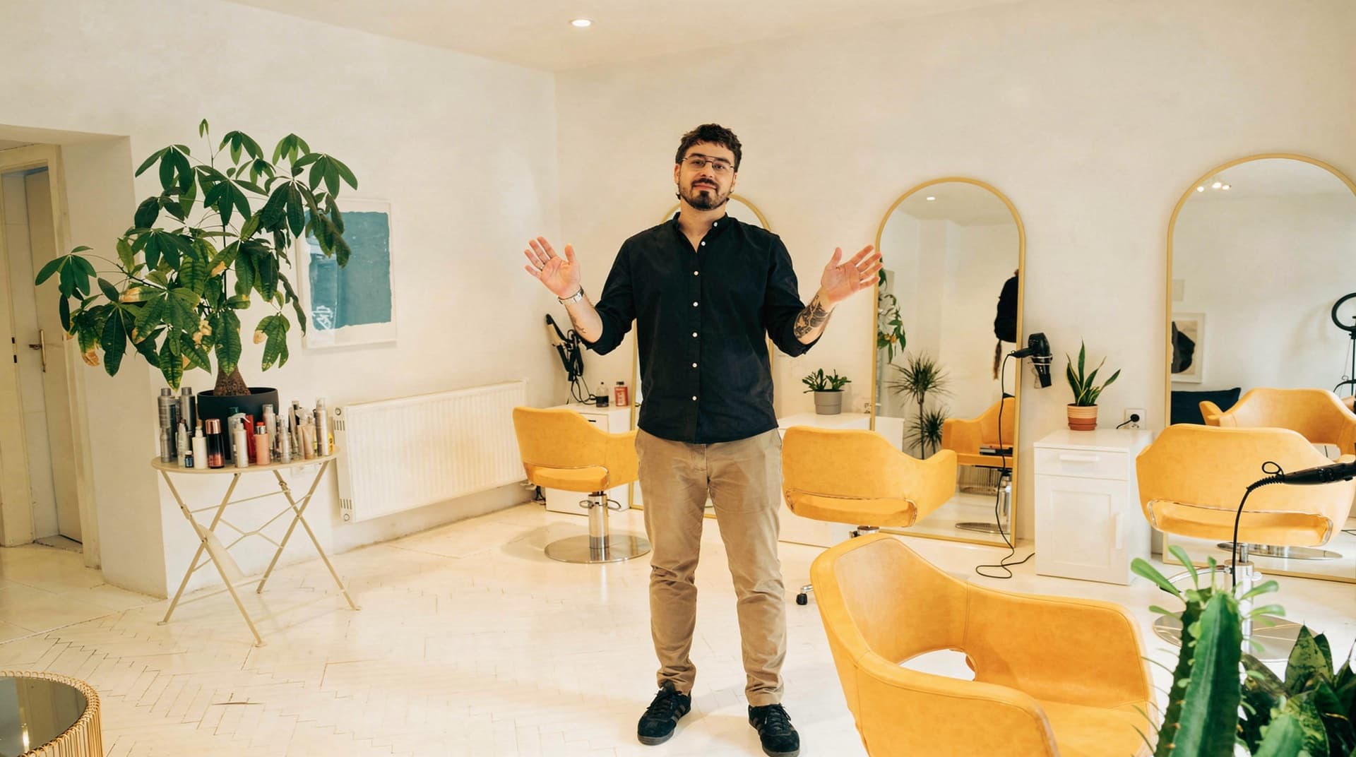 A man stands with raised hands in a salon with yellow chairs and mirrors.