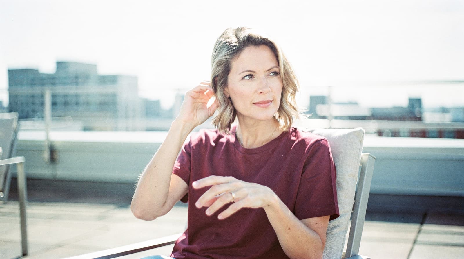 A woman in a red shirt sits on a rooftop patio, looking to the side with buildings in the background.