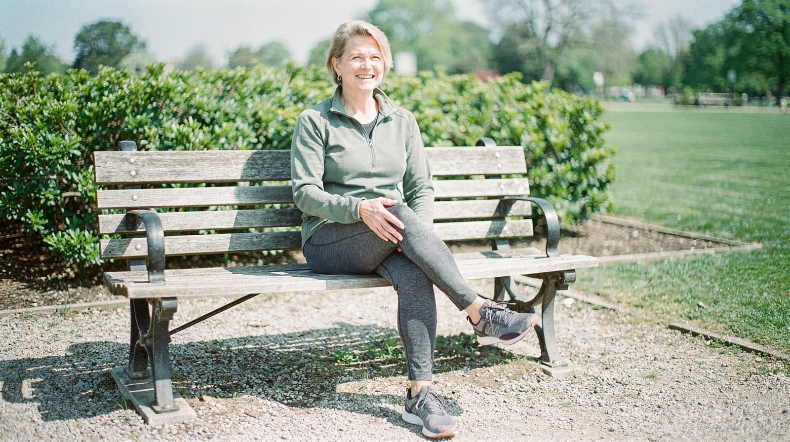A woman sits on a wooden bench in a park, smiling with greenery and a grassy field in the background.
