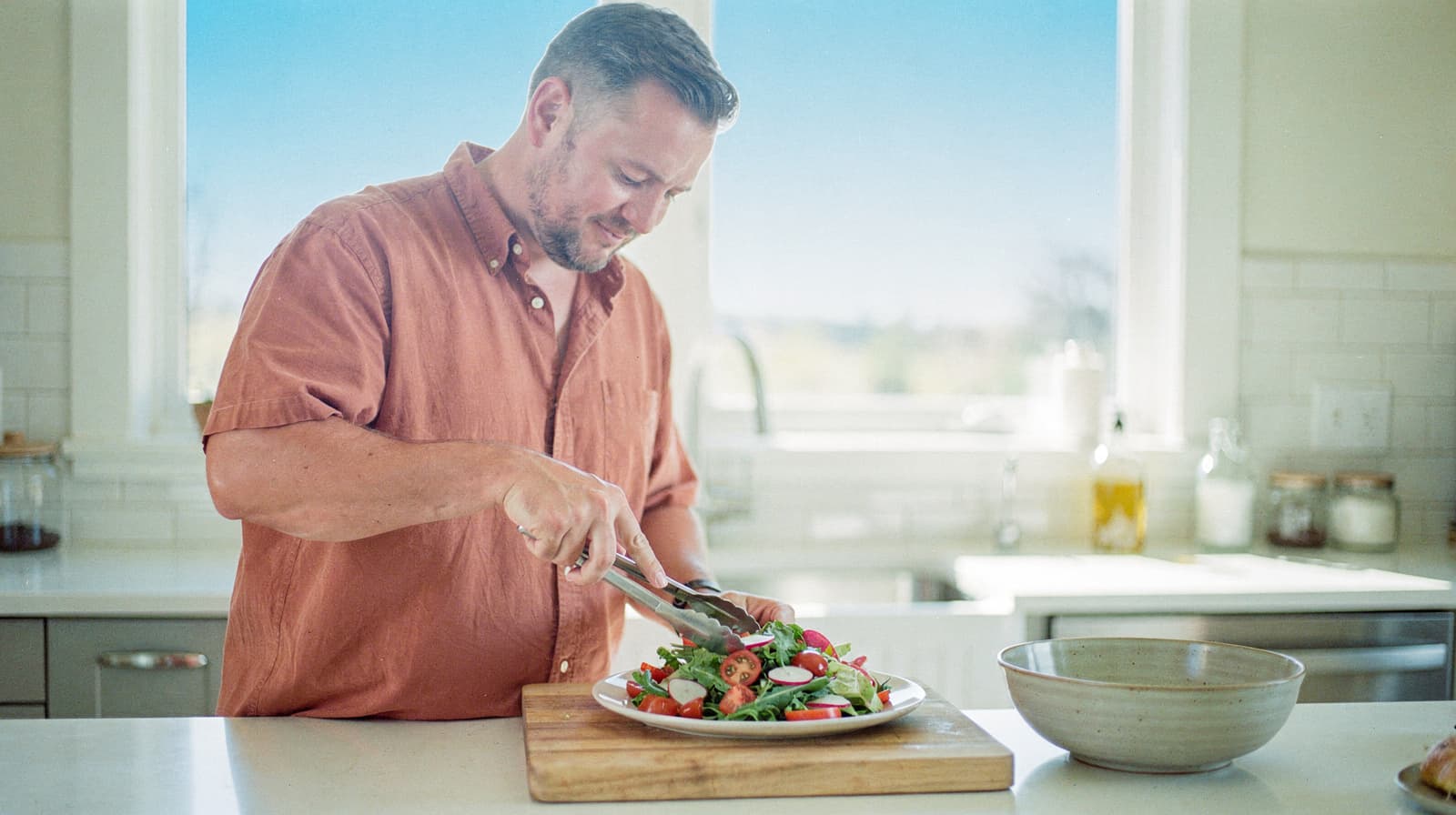 A man in a kitchen prepares a salad with tongs on a wooden cutting board.