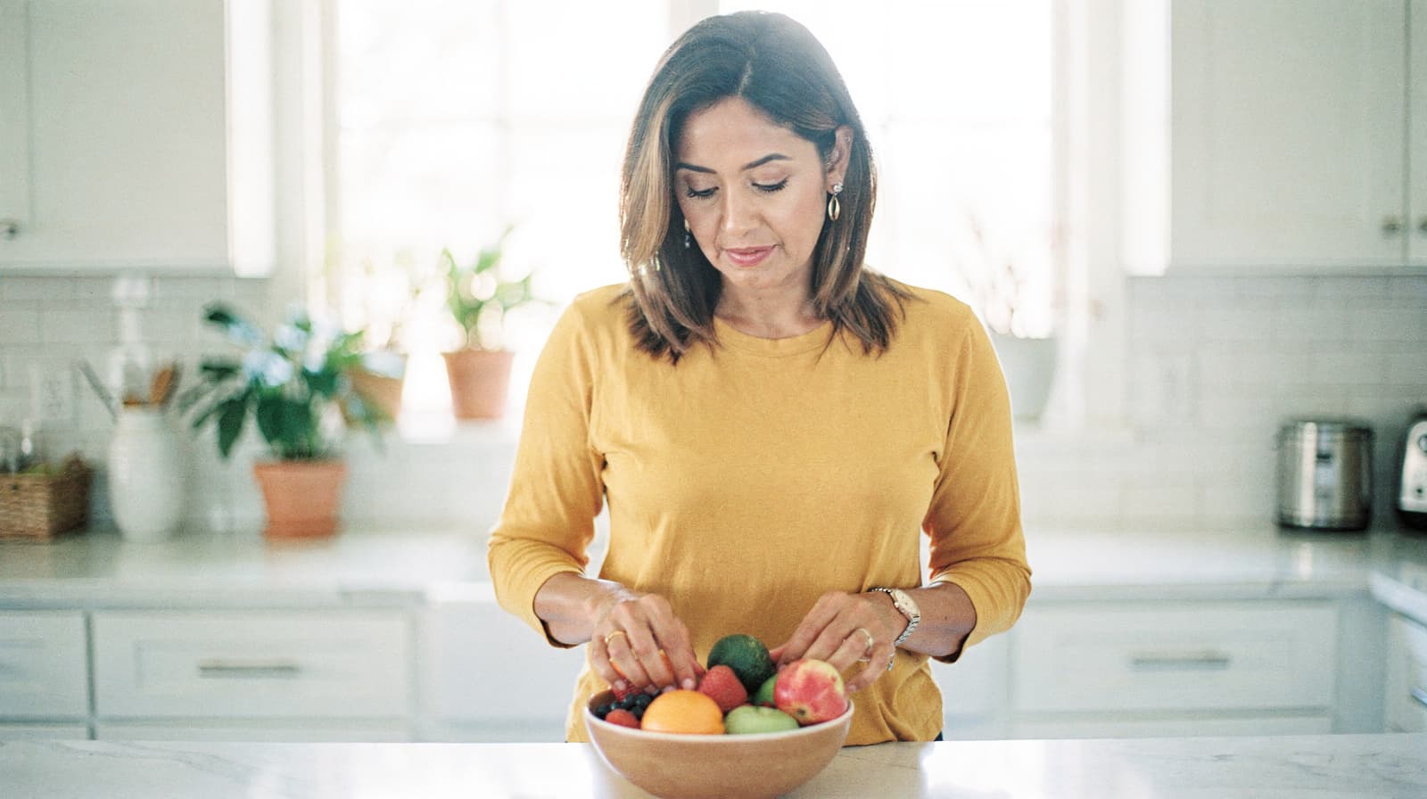A woman in a yellow shirt stands in a kitchen looking at a bowl of fruit on the counter.