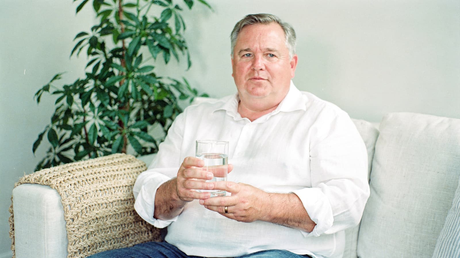 A man in a white shirt sits on a couch holding a glass of water, with a plant in the background.