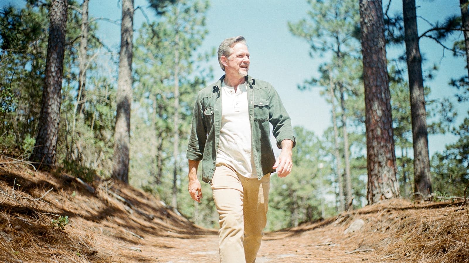 A man walks along a sunlit forest path surrounded by tall pine trees.