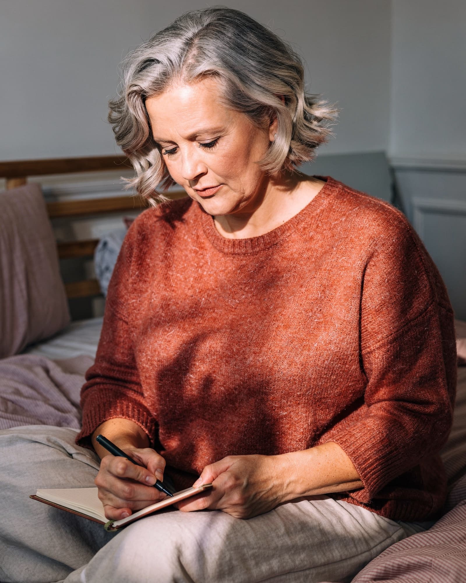 An American woman around fifty journaling calmly on her bed by a bright window, reflective.