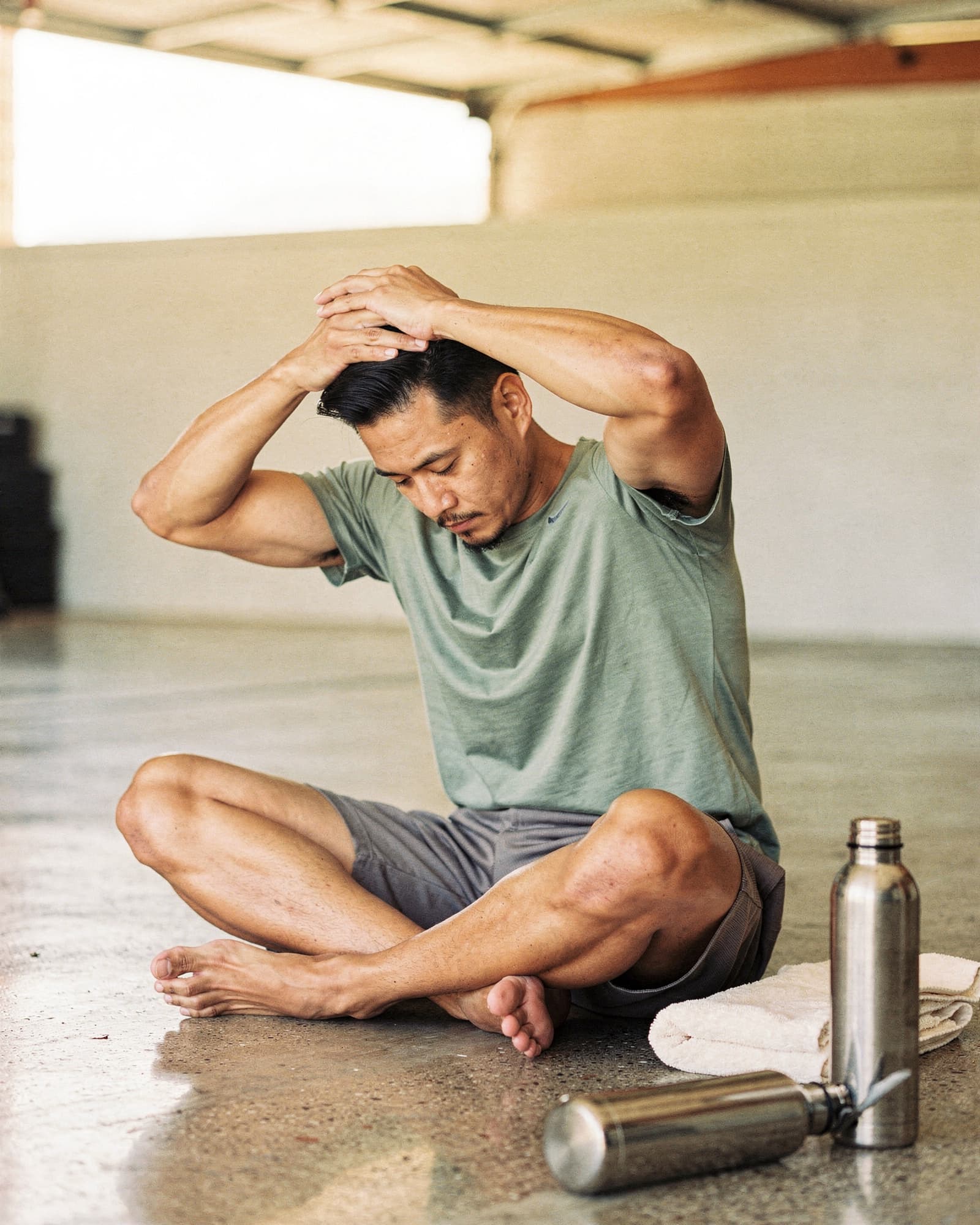 Asian-American man stretching on a sunlit garage floor after training, eyes closed in slow exhale